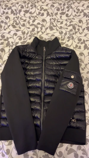 Svart pufferjacka från Moncler Grenoble - Svart pufferjacka från Moncler Grenoble med quiltad front och släta armar. Jackan har ståkrage, två dragkedjefickor och en patch med Moncler-logga på armen. Materialet är polyester och dun, perfekt för kalla dagar. Snygg och sportig look med stilrena detaljer. Kan gå ner i pris vid snabb affär.