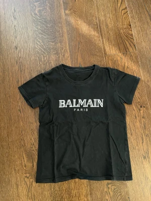 Svart Balmain Paris t-shirt - Svart t-shirt från Balmain Paris med vit logga tryckt framtill. Klassisk rund halsringning och korta ärmar. Perfekt för dig som gillar stilrena och trendiga plagg med exklusiv känsla.