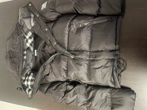 Svart pufferjacka från Burberry - Svart pufferjacka från Burberry London England med rutigt foder i grått och vitt. Jackan har huva, dragkedja och knappar framtill, samt snörning i midjan. Flera fickor med dragkedja och klassisk Burberry-logga på ärmen. Perfekt för kalla dagar.