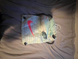 Nike fotbollsskor ljusblå med rosa swoosh - Säljer ett par snygga Nike fotbollsskor i ljusblå färg med rosa swoosh-detaljer. Skorna har snörning och en platt sula med dobbar för bra grepp på planen. Medföljer en Nike-påse med tryck och stor rosa logga. Perfekt för dig som vill sticka ut på fotbollsplanen.