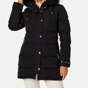 Svart lång pufferjacka med fuskpäls - Svart pufferjacka med lång passform och stor huva med fuskpäls. Jackan har dragkedja och knappar framtill, snörning i huvan och fickor med dragkedja. 