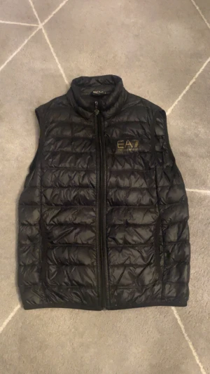  EA7 Emporio Armani väst - Svart äkta quiltad dunväst från EA7 Emporio Armani med guldfärgat logotryck på bröstet. Västen har hög krage och dragkedja framtill med EA7-detalj på zip-pullern. Perfekt att slänga över en hoodie för en clean look.