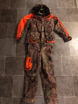 Euro-Hunt camo täckbyxor och jacka  med orange detalj - Säljer ett par täckbyxor och jacka  från Euro-Hunt med camouflagemönster och orange ficka på benet och bröstet. Byxorna har flera praktiska fickor, bälteshällor och är perfekta för utomhusaktiviteter. Materialet är slitstarkt och anpassat för vildmarksliv, 