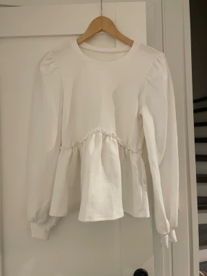 Vit blus med volang och puffärm - Säljer en vit blus med volangdetalj i midjan och puffade axlar. Blusen har rund halsringning och långa ärmar med lätt rynk vid axlarna. Materialet är mjukt och har en lätt struktur, perfekt för dig som gillar en clean och söt look.