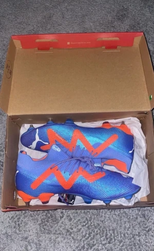 Blå och orange fotbollsskor från Puma - Säljer ett par grymma fotbollsskor från Puma i blått med orange detaljer och vita Puma-loggor. Skorna har snörning och metall-dobbar för extra grepp på planen. Snyggt mönster och modern design, perfekta för dig som vill sticka ut på matchen.