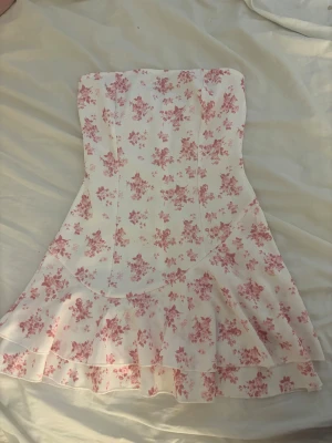 Axelbandslös blommig kortklänning - Säljer en söt axelbandslös kortklänning med volangkant och rosa blommigt mönster på vit botten. Klänningen har en figurnära passform upptill och en lätt utsvängd kjol med dubbla volanger. Perfekt för sommarens alla tillfällen!