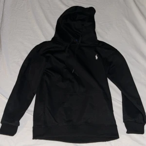 Svart polo ralph lauren hoodie utan zip - Svart hoodie från Polo Ralph Lauren med klassisk vit logga på bröstet. Tröjan har huva med snörning, stor magficka och ribbade muddar vid ärmslut och nederkant. Tillverkad i mjuk bomull, perfekt för chill dagar. Snygg och enkel design som alltid funkar. storlek M men passar mer som S