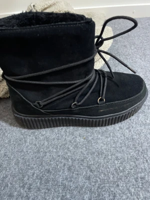 Svarta moon boots från Everest - Moon boots från Everest i svart mocka med snörning runt hela skaftet och grov platt sula. Insidan är fodrad för extra värme och komfort. Perfekt för kalla vinterdagar och har en modern, chunky look som sticker ut.