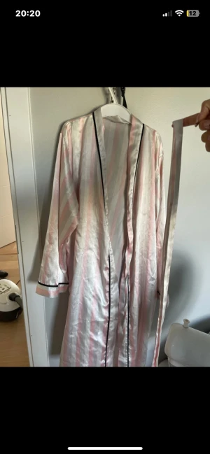 Randig lång morgonrock i satin - Säljer en lång morgonrock i glansig satin med ljusrosa och vita ränder. Kimonon har svarta kantdetaljer och medföljer ett matchande bälte att knyta i midjan. Perfekt för en chill vibe hemma eller när du vill känna dig extra lyxig.