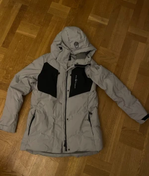Grå Sail Racing pufferjacka med huva - Säljer den perfekta gråa vinterjackan från Sail Racing med svarta detaljer och logga på bröstet och huvan. Jackan har praktiska fickor med dragkedja. Perfekt för kalla dagar och har en snygg och sportig look. Storlek S/Small men passar även M/medium. Inga defekter! Säljer den eftersom den tyvärr blivit för liten för mig.