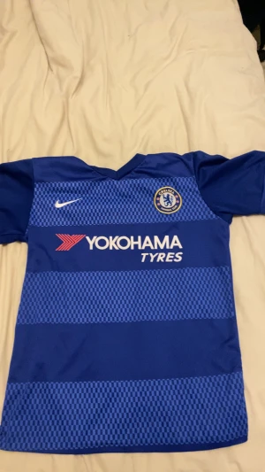 Chelsea FC Matic 21 - Chelsea FC fotbollströja med Matic och nummer 21 på ryggen. Blå med vita detaljer, randigt rutmönster på framsidan, klubbmärke och Nike-logga. Materialet är lätt och andas, perfekt för träning eller match. Sponsorn Yokohama Tyres tryckt på bröstet.