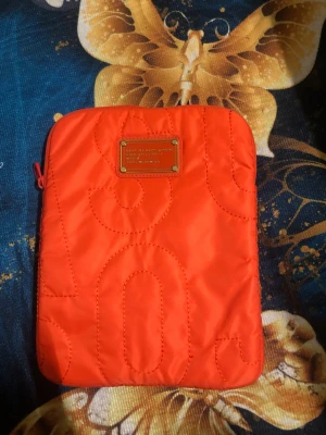 Orange kuvertväska Marc by Marc Jacobs - Säljer en snygg orange iPadfodral från Marc by Marc Jacobs med quiltad design och metallplakett framtill. Väskan är gjord i mjuk polyester och har en dragkedja med coolt ringformat zip-tag. Perfekt för att hålla koll på dina essentials.