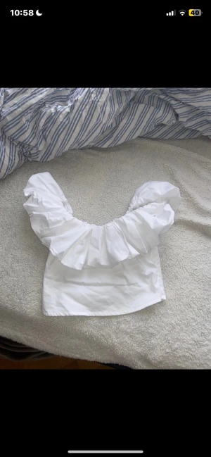 Vit volang topp från Amisu - Supersöt vit topp från Amisu med breda volanger över axlarna och offshoulder-design. Croppad modell med dragkedja i sidan för enkel påklädning. Gjord i mjukt syntetmaterial som känns skönt mot huden. Perfekt för dig som gillar statement-plagg och vill sticka ut.