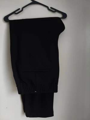 Svarta dressade byxor från Zara XL - Svarta dressade byxor från Zara i storlek XL med elastisk midja och knäppning fram. Byxorna har en rak passform, sidfickor och är tillverkade i ett mjukt material som ger en clean och stilren look. Perfekta för dig som gillar enkel och snygg stil. Eleganta men väldigt bekväma.