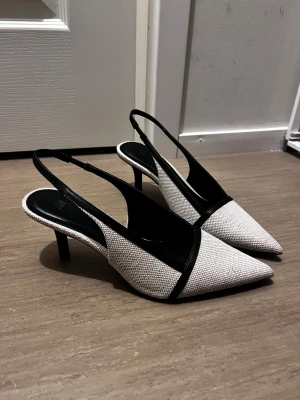 Svarta och vita pumps med spetsig tå - Snygga pumps från H&M med spetsig tå och låg klack. Skorna är vita med svart kant och rem, tillverkade i textil med struktur och syntetmaterial. Perfekt för dig som gillar stilrena och trendiga skor med en elegant touch.