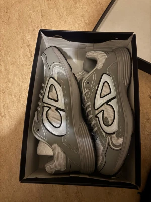 Grå Dior sneakers med CD-detalj - Snygga grå sneakers från Dior med tydlig CD-logga på sidan. Skorna har en rund tå, platt sula och är tillverkade i en mix av syntet och mesh för en modern look. Silverfärgade detaljer och snörning framtill ger en clean och trendig vibe.