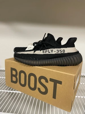 Adidas Yeezy Boost 350 V2 Black/White - Adidas Yeezy Boost 350 V2 i svart och vit med SPLY-350 på sidan. Skorna har en stickad ovandel i syntet, platt sula med boost-dämpning och snörning. Ikonisk siluett med rund tå och cool kontraststripe. Perfekt för dig som vill ha streetstyle med hype.