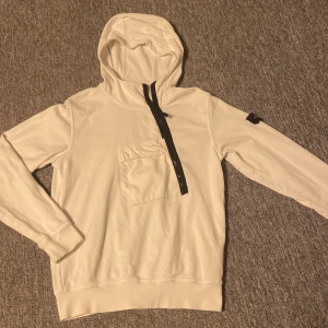 Vit hoodie från Stone Island  - Vit hoodie från Stone Island Junior med svart half zip framtill och klassisk logotyp-patch på vänster ärm. Tröjan har en stor ficka på bröstet, huva och långa ärmar. Tillverkad i mjuk bomull, perfekt för chill dagar. I tröjan står de bara junior men skulle säga att de sitter ungefär som XS i herr storlekar. Köpt för ca 3000. Används ganska flitigt men fortfarande i bra skick. OBS lite hål under logan på höger armen(se bild 5) men inget man märker. Skriv vid minsta fundering.