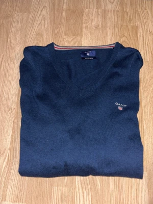 Marinblå v-ringad tröja från GANT - Snygg marinblå v-ringad tröja från GANT i storlek L men passar bättre M. Tröjan är tillverkad i en mjuk blandning av bomull och ull, har långa ärmar och ribbade muddar. Diskret GANT-logo broderad på bröstet och en cool randig detalj i kragen.