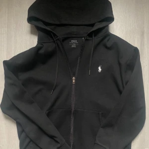 Svart hoodie från Polo Ralph Lauren - Svart hoodie från Polo Ralph Lauren med dragkedja och klassisk vit logga på bröstet. Tröjan har huva med snörning och känguruficka framtill. Perfekt för chill dagar och enkel att matcha med allt. Materialet är mjuk bomull för extra komfort. (inte mina bilder)