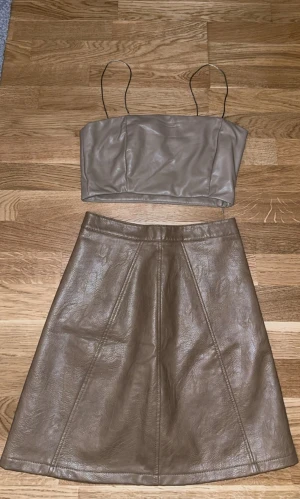 Set - Crop Top och Kjol från Zara - Snygg beige kortkjol från Zara Basic Collection i skinnimitation. Kjolen har en A-linjeformad siluett med dekorativa sömmar och dragkedja framtill med dubbla knappar i midjan. Perfekt för dig som gillar stilrena och trendiga plagg.