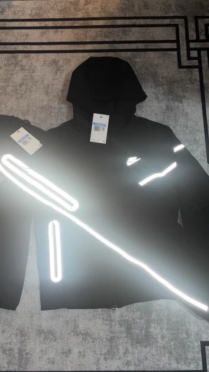 Nike svart träningsset med reflex - Nike svart träningsset med hoodie och matchande byxor. Snygga vita reflexdetaljer på armar, bröst och ben som lyser upp i mörker. Setet har dragkedja på jackan och fickor, samt Nike-logga på bröstet. Där du kan säkra att den är äkta o tar dig till Nike hemsidan. Perfekt för dig som vill sticka ut när du tränar utomhus.