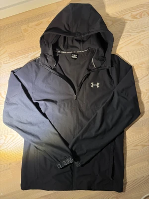 Svart vindjacka från Under Armour - Svart vindjacka från Under Armour med huva och dragkedja hela vägen. Jackan har snygga detaljer med logga på bröstet och snörning i huvan. Perfekt för träning eller när du vill ha en sportig look. Materialet är lätt och syntetiskt, vilket gör den smidig att bära.