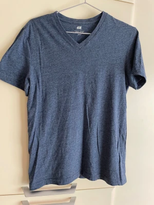 Blå v-ringad t-shirt från H&M - Snygg blå t-shirt från H&M med v-ringad hals och regular fit. T-shirten är kortärmad och tillverkad i mjuk bomull, perfekt för en chill och enkel stil. Passar dig som gillar klassiska och bekväma plagg med en modern touch.