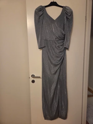 Glittrig silver långklänning med puffärm - Snygg långklänning i glittrigt silver med puffade axlar och v-ringad omlottfront. Klänningen har trekvartsärmar och ett elegant fall med drapering framtill. Perfekt för dig som vill sticka ut med en glamorös look. Dragkedja i ryggen för enkel påklädning.