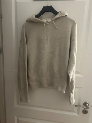 Beige stickad hoodie med huva - Mysig beige stickad hoodie med huva och snörning. Tröjan har långärmade ärmar och ribbad nederkant som ger en chill och avslappnad vibe. Perfekt för att hålla sig varm under kyliga dagar. Enkel och stilren design som passar till det mesta.