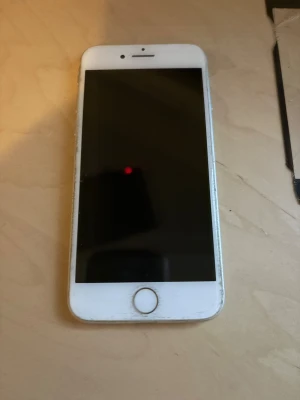 iPhone 8 - Apple iPhone SE i vitt utförande. Skärmen och kanterna visar tydligt slitage och repor, men enheten är hel. Touch ID och kamera synliga. Perfekt för dig som söker en prisvärd iPhone med klassisk design och kompakt format.