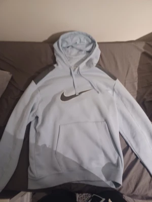 Ljusblå Nike hoodie med stor logga - Nike hoodie i ljusblått med stor svart och vit swoosh på bröstet. Tröjan har huva med snörning, magficka och långa ärmar. Mjuk insida och klassisk passform, perfekt för chill eller träning. Snygga grå detaljer på axlarna ger extra stil.