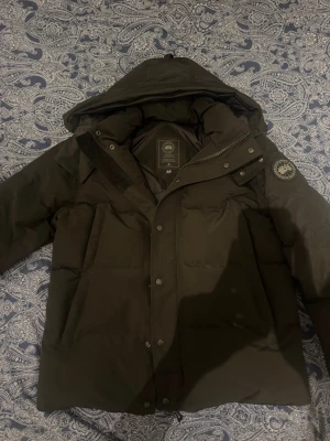 Svart dunjacka från Canada Goose blacke label  - Svart dunjacka från Canada Goose med huva och klassisk svart patch på ärmen. Jackan har dragkedja och knappar framtill, samt stora fickor. Materialet är slitstark polyester och dunfyllning som håller dig varm under kalla dagar. Perfekt för dig som vill ha en stilren och funktionell vinterjacka.