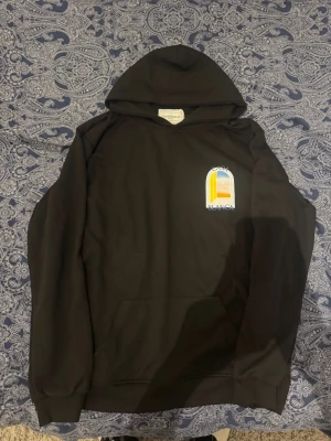 Svart hoodie Casablanca Tennis Club L - Svart hoodie från Casablanca Tennis Club i storlek L. Framsidan har ett litet tryck med färgglad grafik och texten 'Casa Blanca', medan ryggen har ett stort grafiskt tryck i blått, gult och orange. Klassisk huva och magficka, perfekt för en chill look.