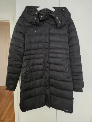 Svart dunjacka Calvin Klein Jeans M - Svart lång dunjacka från Calvin Klein Jeans i storlek M. Jackan har quiltad design, huva med tryckknappar, dragkedja och ribbade muddar vid ärmslut. Fylld med dun och fjäder för extra värme. Snygg och stilren look med praktiska fickor. Päls till luva saknas. 