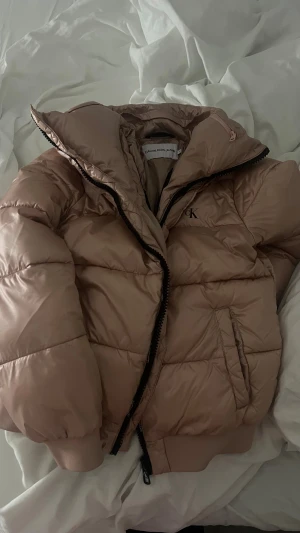 Beige vinterjacka från Calvin Klein - Snygg beige pufferjacka från Calvin Klein Jeans med svart dragkedja och CK-logga på bröstet. Jackan har två sidofickor och är tillverkad i lätt, syntetiskt material med Dupont Sorona-fyllning för extra värme. Har även en inbyggd luva i kragen. Pris kan sänkas vid snabb affär. Nypris runt 3000 kr.