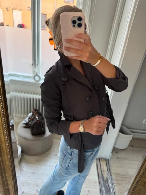 Svart kort trenchcoat med bälte - Säljer en svart, kort trenchcoat med dubbelknäppning och bälte i midjan. Jackan har klassisk krage, långa ärmar och snygga axelremmar med knappar. Perfekt för dig som vill ha en stilren och tidlös look.