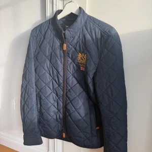 Mörkblå quiltad jacka från Morris - Quiltad mörkblå jacka från Morris med snyggt broderat emblem och brittisk flagga på bröstet. Jackan har ståkrage, dragkedja framtill och diskreta fickor. Perfekt för dig som gillar klassisk stil med modern touch.