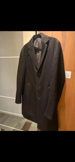 Brun rock i ull med klassisk krage - Snygg brun rock med klassisk krage och två knappar framtill. Jackan har två fickor på sidorna och är helfodrad på insidan. Materialet är mjuk ull som ger en stilren och clean look. Perfekt för dig som gillar minimalistisk stil och vill ha en tidlös jacka.