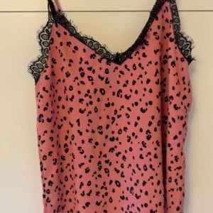 Rosa leopardmönstrad linnetopp med spets - Säljer en rosa linnetopp med svart leopardmönster och snygga svarta spetsdetaljer vid halsringningen och axelbanden. Toppen är ärmlös och har en v-ringad form som ger en trendig vibe. Perfekt för dig som gillar djurmönster och coola detaljer.