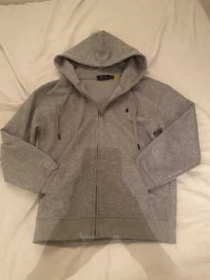 Grå hoodie från Polo Ralph Lauren - Snygg grå hoodie från Polo Ralph Lauren med dragkedja och luva. Använts 2-3 gånger nästan helt ny.