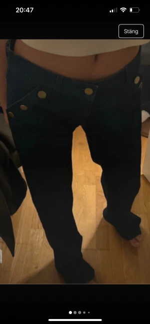 Svarta wide jeans med guldknappar - Snygga svarta jeans med låg midja och breda ben. Byxorna har flera guldiga knappar framtill som cool detalj och klassisk femficksdesign. Passar dig som gillar en loose och avslappnad stil. Materialet är jeans och färgen är djup svart.