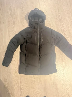 Svart Sail Racing dunjacka med huva - Svart dunjacka från Sail Racing, modell Antarctica Expedition. Jackan har huva, två bröstfickor med dragkedja och Sail Racing-logga på bröstet. Tjockt, quiltat material som håller värmen och stilren design. Perfekt för kalla vinterdagar och äventyr utomhus. Den är i storlek 170 men passar mig jag är 177