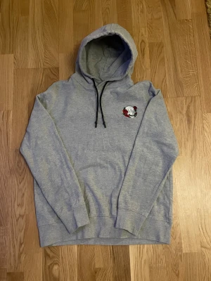 Grå hoodie med döskalle och rosor - Snygg grå hoodie från The Black Label Collection med broderad döskalle och röda rosor på bröstet och ett stort tryck på ryggen med texten 'Live Life No Regrets'. Tröjan har huva med svarta snören och är långärmad. Perfekt för dig som gillar edgy streetstyle. Storlek L men passar mer som en M.