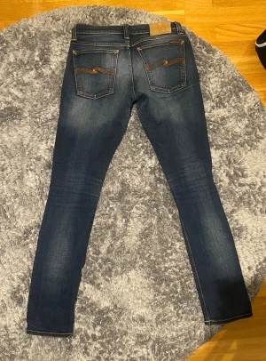 Mörkblå Nudie Jeans - Modell Grim Tim och i princip helt nya. Snygga mörkblå jeans från Nudie Jeans slim fit och klassisk. Jeansen har orangea sömmar och coola broderade detaljer på bakfickorna. Tillverkade i slitstarkt denim med lätt tvättad look. Perfekt för dig som gillar stilrena och lättmatchade jeans.