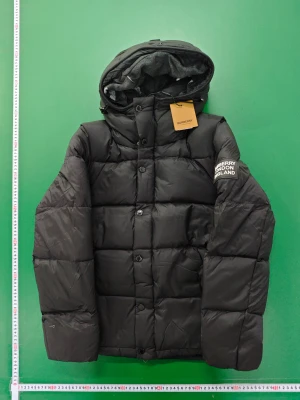 Svart pufferjacka från Burberry M - Svart pufferjacka från Burberry med huva och quiltad design. Jackan har tryckknappar framtill och logotypen 'BURBERRY LONDON ENGLAND' på vänster ärm. Insidan av huvan har ett rutigt mönster i grått och rött. Perfekt för kalla dagar med sin oversized look.