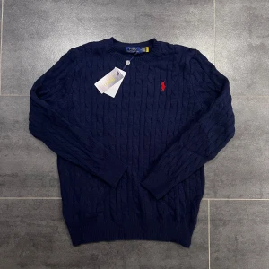 Mörkblå stickad tröja från Polo Ralph Lauren - Snygg mörkblå stickad tröja från Polo Ralph Lauren med klassiskt kabelstickat mönster och rund hals. Tröjan har långärmade ärmar och en röd broderad logga på bröstet. Perfekt för dig som gillar stilrena och tidlösa plagg.