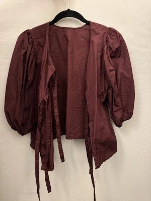 Vinröd topp med puffärmar - Säljer en vinröd topp med puffiga långärmar och långa knytband framtill. Toppen har en loose passform och är tillverkad i bomull. Perfekt för dig som gillar statement-ärmar och vill ha en cool detalj till din outfit.