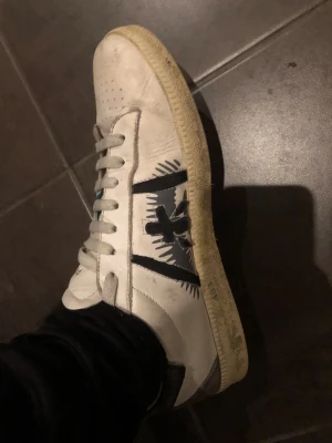 Premiata vita sneakers med svart print - Säljer ett par vita sneakers från Premiata med coolt svart och grått grafiskt print på sidan. Skorna har snörning, perforerad tå och beige sula. Materialet är skinn och detaljerna ger en unik vibe. Perfekt för dig som gillar streetstyle och vill sticka ut.
