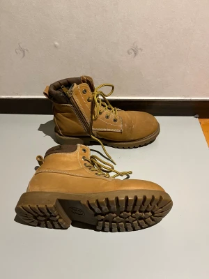 Brungula kängor - Säljer ett par robusta boots i beige skinn med grov sula och rund tå, väldigt lika Timberland kängorna. Skorna har gulbruna snören, vit kontrastsöm och dragkedja på sidan för enkel påtagning, snörningen och dragkedjan är i utmärkt skick. Mjukt vadderad brun krage upptill för extra komfort. Perfekt för höst och vinter. Finns tecken på användning under hälen men inget som påverkar användningen